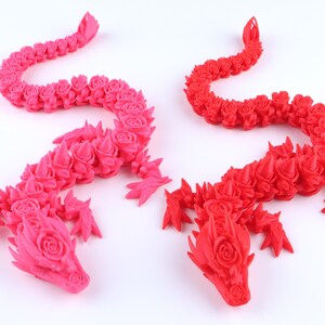 May include: Two articulated dragon figurines, one in bright pink and one in vibrant red. Each dragon features a detailed, segmented body composed of repeating rose-like floral patterns, with sharp, pointed spikes along the back and head.