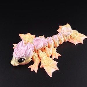 May include: A whimsical, pastel-colored dragon figurine with a floral design. The dragon's body is a gradient of yellow and orange, with pink rose accents on its head, back, and tail. The figurine is set against a black background.