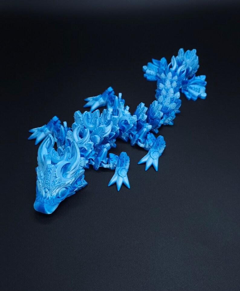 May include: A bright blue articulated lizard figurine featuring a complex, layered scale pattern across its body, head, and tail. The creature has four legs with pointed claws and a detailed, textured surface design throughout its entire form.
