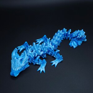 May include: A bright blue articulated lizard figurine featuring a complex, layered scale pattern across its entire body. The creature has a detailed head with prominent horns, four clawed feet, and a long, segmented tail that curves slightly.
