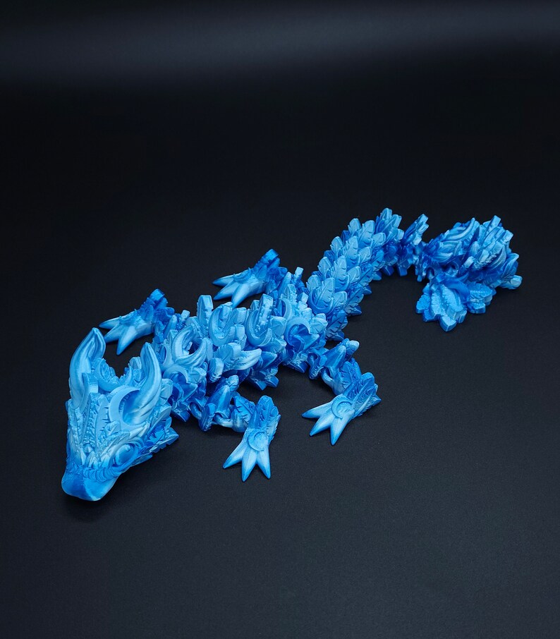 May include: A bright blue articulated lizard figurine featuring a complex, layered scale pattern across its entire body. The creature has a detailed head with prominent horns, four clawed feet, and a long, segmented tail that curves slightly.