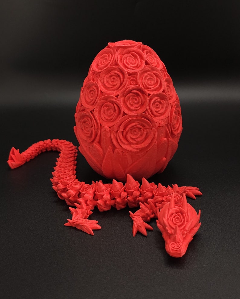 May include: A bright red 3D printed articulated dragon figure with a rose-textured body, positioned next to a matching egg-shaped container covered in a detailed pattern of sculpted roses, both featuring a matte finish and intricate floral details.
