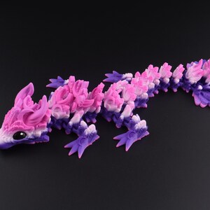 May include: An articulated dragon figurine featuring a gradient color scheme that transitions from deep purple at the base to bright pink and white along the dorsal ridges. The creature has large black eyes and a segmented body with four legs.