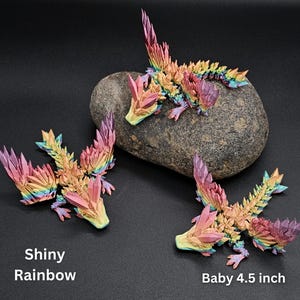 Puede incluir: Tres figuras de drag&oacute;n articuladas con un acabado brillante de arco&iacute;ris y texturas cristalinas. Un drag&oacute;n descansa sobre una piedra gris, mientras que otros dos est&aacute;n colocados cerca. El texto en la imagen dice Shiny Rainbow y Baby 4.5 inch.