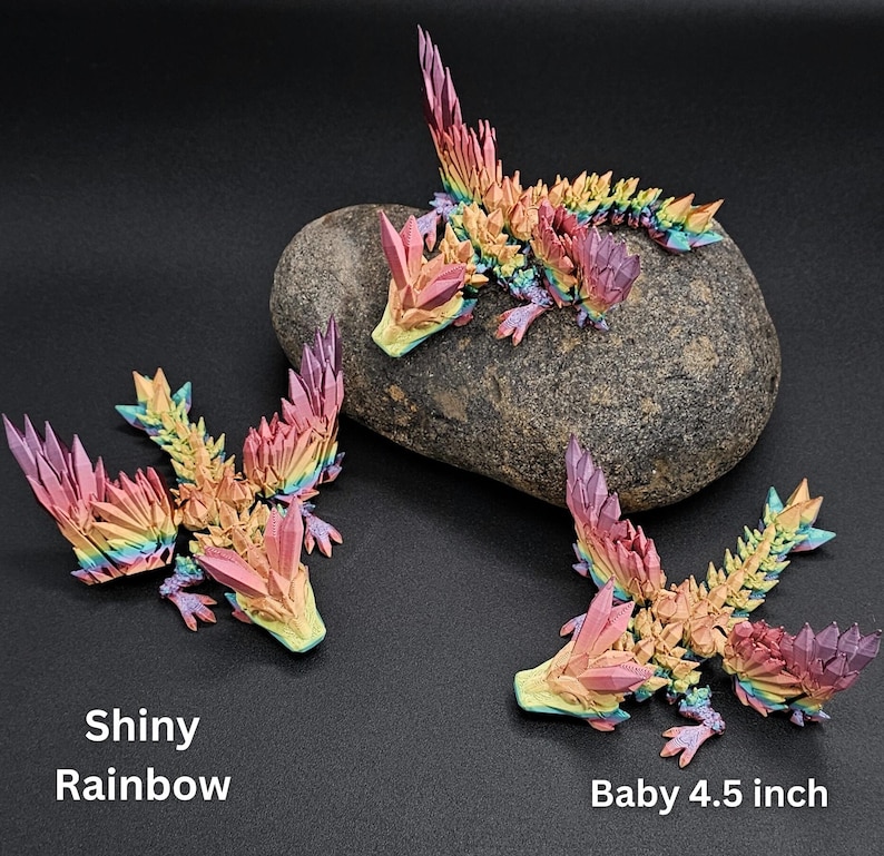 Puede incluir: Tres figuras de drag&oacute;n articuladas con un acabado brillante de arco&iacute;ris y texturas cristalinas. Un drag&oacute;n descansa sobre una piedra gris, mientras que otros dos est&aacute;n colocados cerca. El texto en la imagen dice Shiny Rainbow y Baby 4.5 inch.