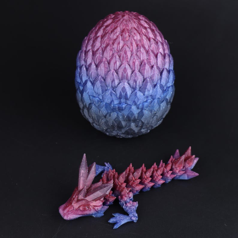 May include: A 3D-printed dragon and egg set. The egg is a gradient of pink, purple, and blue, with a textured, scale-like pattern. The dragon is in a similar color scheme, with crystal-like spikes along its back and head. The background is black.