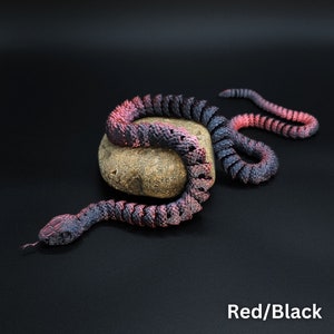 May include: A red and black rubber snake coiled around a rock. The snake is made of a flexible material and has a realistic texture.