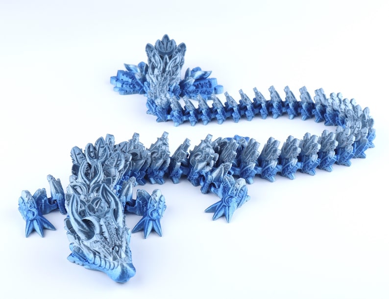 May include: A 3D printed articulated dragon figurine featuring a metallic blue and silver gradient finish. The creature has a detailed head with horn-like protrusions, four clawed feet, and a long, segmented body that curves across the frame.