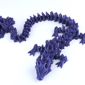 May include: A dark purple articulated dragon figurine with a segmented body, detailed scales, and sharp claws. The creature features a long, winding tail and a head with prominent horns, showcasing a complex, layered design throughout its entire form.
