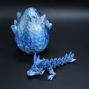 May include: A blue, crystal-like dragon and egg set. The egg is open, revealing a detailed interior. The dragon is in a crawling position, with crystal-like spikes along its back and head. The set is made of a light blue material.