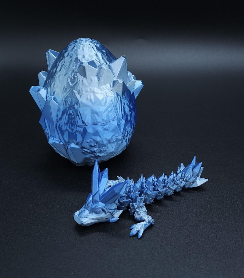 May include: A blue, crystal-like dragon and egg set. The egg is open, revealing a detailed interior. The dragon is in a crawling position, with crystal-like spikes along its back and head. The set is made of a light blue material.