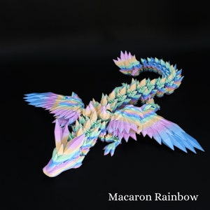Puede incluir: Una figura de drag&oacute;n articulada multicolor con un degradado de arco&iacute;ris pastel en rosa, amarillo, azul y verde. La criatura tiene escamas, alas y una cola larga de forma geom&eacute;trica y cristalina. El texto Macaron Rainbow est&aacute; impreso en blanco.