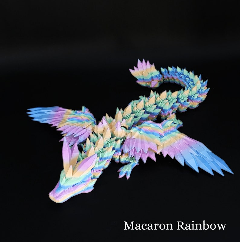 Puede incluir: Una figura de drag&oacute;n articulada multicolor con un degradado de arco&iacute;ris pastel en rosa, amarillo, azul y verde. La criatura tiene escamas, alas y una cola larga de forma geom&eacute;trica y cristalina. El texto Macaron Rainbow est&aacute; impreso en blanco.