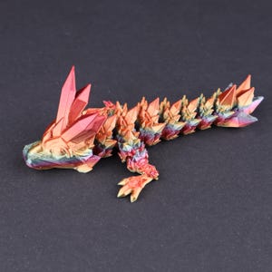 May include: A colorful, articulated dragon figurine with a crystal-like design. The dragon is primarily orange, red, and gold, with hints of blue and green. The figurine has a detailed, textured surface and is posed on a dark background.