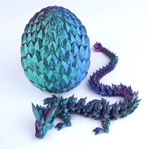 May include: An iridescent, multi-colored dragon figurine with a crystalline, scale-like texture in shades of deep blue, purple, and teal, positioned next to a matching, large, oval-shaped egg with a similar faceted, scale-like surface pattern.