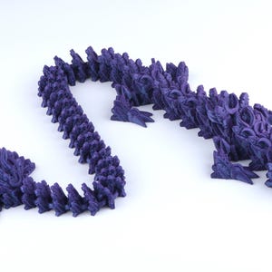 May include: A dark purple articulated dragon figurine featuring a segmented body with intricate scale textures, a detailed head with horns, and four limbs. The flexible design allows the creature to curve into various positions on a plain surface.