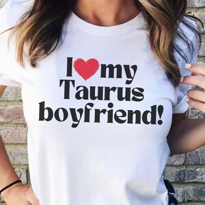 May include: White t-shirt with black text that reads "I love my Taurus boyfriend!" with a red heart replacing the "o" in love.