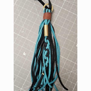 May include: A decorative tassel made of black and turquoise leather strips. The tassel features a braided leather center, a brown leather wrap at the top, a gold-toned metal ring, and a gold-toned metal tube accent. A black leather cord loop is at the top.