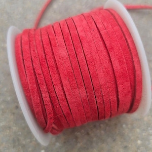Op de afbeelding: Een spoel helderrood suède koord. Het koord is strak gewikkeld rond een witte plastic spoel. Het koord lijkt plat en heeft een levendige, effen rode kleur. De achtergrond is een neutraal grijs.