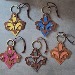 May include: Five leather keychains in a fleur-de-lis design. Each keychain features a different color scheme: yellow, blue, pink, gray, and orange. They have a brown leather strap and a metal ring for attaching keys.