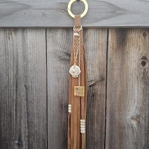 Puede incluir: Un llavero de borla de cuero marrón con un anillo dorado y un pequeño dije. El llavero presenta una concha, una cadena dorada y un dije cuadrado con texto. Cuentas blancas están ensartadas a lo largo de la borla.