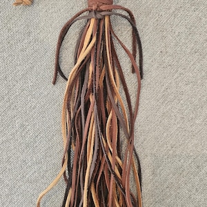 May include: A leather tassel with a bronze ring and a loop. The tassel is made of multiple strands of leather in various shades of brown, tan, and black. The loop is made of a lighter brown leather.