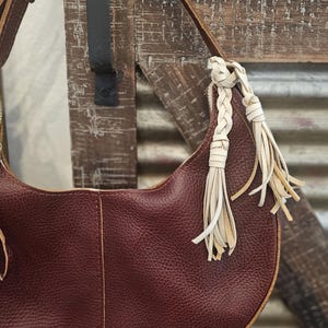 May include: A burgundy leather shoulder bag with a crescent shape. The bag features a long, adjustable strap and decorative white leather tassels. The bag is hanging on a wooden surface.