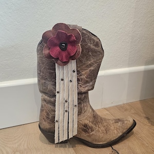 May include: A brown leather cowboy boot with a distressed finish, decorated with a burgundy flower and white fringe. The boot has a pointed toe and a floral accent, suitable for Western-style attire.