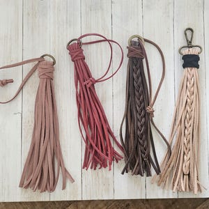 May include: Four leather tassels in dusty rose, deep magenta, dark brown, and beige. Each tassel features thin leather strips, a knotted or braided top section, and a metal ring or lobster clasp attachment for hanging or securing to items.
