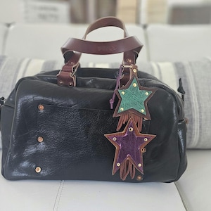 May include: Black leather handbag with a top handle and decorative star-shaped charms. The bag features a textured surface, brass rivets, and a brown leather handle. The star charms are green and purple with gold accents and fringe.