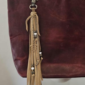 May include: A burgundy leather tote bag with a decorative tassel. The tassel is made of tan suede strips, gold-colored chains, beads, and leaf charms. The bag has a black leather strap and a metal clasp.