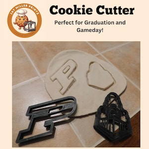 May include: Black plastic cookie cutters shaped like a letter P and a graduation cap. The cutters are on a light brown tiled surface with a white dough imprint of the letter P and the graduation cap.