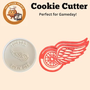 Könnte beinhalten: Roter und weißer Ausstecher in Form des Detroit Red Wings Logos. Das Logo zeigt einen roten Flügel mit einem weißen Rad im Inneren. Der Ausstecher ist aus rotem Kunststoff.