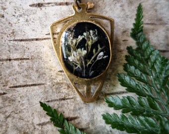 May include: A brass-coloured pendant necklace. The pendant has a shield-like shape and features a circular charm filled with resin and dried white flowers set against a black backdrop. The necklace is displayed on a light-coloured wooden surface, with green fern leaves.