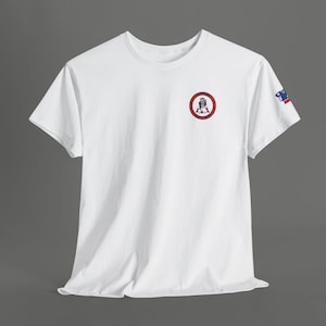 May include: White t-shirt with a crew neck and short sleeves. A circular emblem featuring a figure in a tricorn hat is on the chest. A small flag design is on the left sleeve. The shirt is made of a soft material.