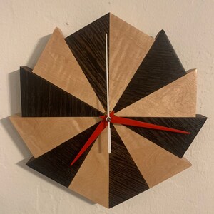 May include: A geometric wooden clock with a red hour hand and a white minute hand. The clock face is made of alternating dark wood and light wood triangles.