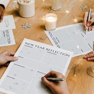 Op de afbeelding: Witte vellen papier met de titel NEW YEAR REFLECTION met secties voor prestaties, uitdagingen en doelen. De vellen hebben een lichtgrijs sneeuwvlokpatroon en liggen op een houten tafel met pennen, een kaars en glazen.