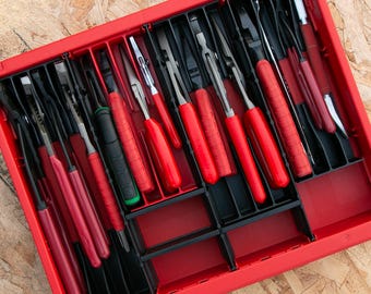 May include: A red plastic tool organizer tray featuring multiple vertical slots containing various hand tools with red and black handles, including pliers and cutters, arranged in a structured grid for storage and organization.