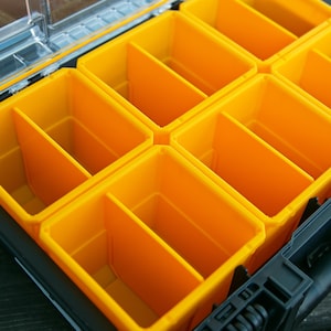 Peut inclure: Une mallette de rangement en plastique noir dotée de plusieurs bacs amovibles jaune vif. Chaque bac jaune est divisé en deux sections par une paroi verticale centrale. La mallette comprend un couvercle transparent à charnière visible en haut.