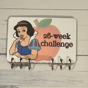 May include: A white and red 26-week challenge planner with a cartoon image of Snow White and a red apple. The planner has a silver ring binding.