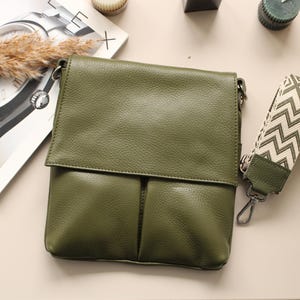 Peut inclure: Sac bandouli&egrave;re en cuir vert olive avec fermeture &agrave; rabat et bandouli&egrave;re &agrave; motifs. Le sac a un aspect textur&eacute; et un fermoir argent&eacute;. Un magazine et d'autres accessoires sont en arri&egrave;re-plan.