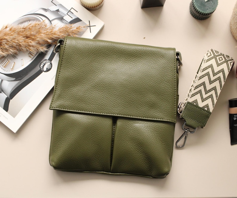 Peut inclure: Sac bandouli&egrave;re en cuir vert olive avec fermeture &agrave; rabat et bandouli&egrave;re &agrave; motifs. Le sac a un aspect textur&eacute; et un fermoir argent&eacute;. Un magazine et d'autres accessoires sont en arri&egrave;re-plan.