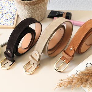 Puede incluir: Tres cinturones de ante en marrón, beige y marrón claro, cada uno con una hebilla dorada. Los cinturones están enrollados, mostrando su forro interior de cuero. La imagen también incluye productos de belleza y una planta decorativa.