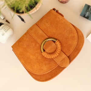 May include: A tan suede shoulder bag with a rounded flap and a decorative circular clasp. The bag has a stitched design and a strap. The bag is on a white surface with other objects, including a small candle and a magazine.