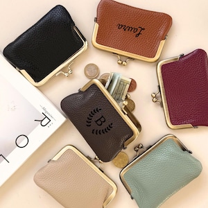 May include: A collection of leather coin purses in colours such as black, brown, and burgundy, with gold-toned clasps. One purse is open, showing coins and banknotes. Some purses have personalized text or a monogram.