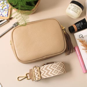 May include: A beige leather crossbody bag with a patterned strap, a gold-tone clasp, and a tassel. The bag is surrounded by beauty products, a magazine, and a pen.
