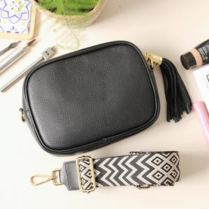 May include: Black crossbody bag with a detachable patterned strap. The bag features a gold-tone zipper pull with a black tassel. A tube of foundation, a pink tube of mascara, and a magazine are also in the image.