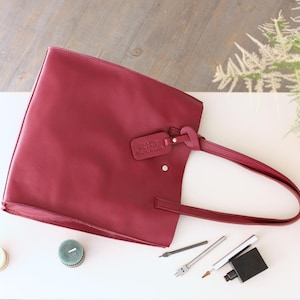 Könnte beinhalten: Eine bordeauxfarbene Ledertasche mit passendem Griff und kleinem Anhänger. Die Tasche ist rechteckig und liegt auf einer weißen Oberfläche. Verschiedene Bastelwerkzeuge und Kerzen sind um die Tasche herum angeordnet.