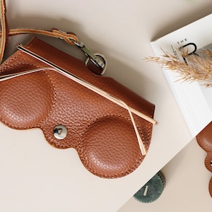 May include: A brown leather eyeglasses case with a strap and a silver clasp. The case has a unique shape with two rounded sections and a snap closure. A pair of eyeglasses rests inside the case. A magazine and dried plants are in the background.