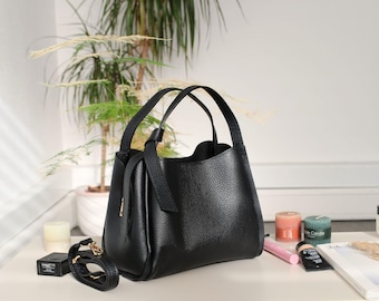 May include: A black leather handbag with a textured finish, featuring two top handles and a detachable shoulder strap. The bag is set on a white surface with beauty products and a plant in the background.